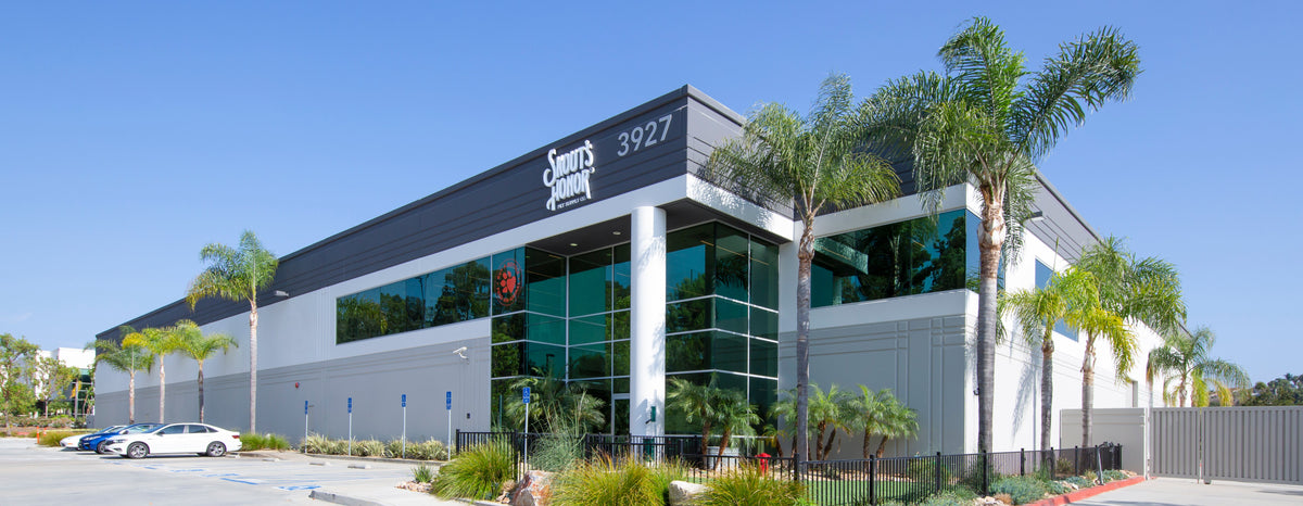 A modern commercial building with a dark gray roof, white and gray facade, large glass windows, and palm trees in front, under a clear blue sky. The building signage reads 'Skout's Honor HQ' and '3927'.